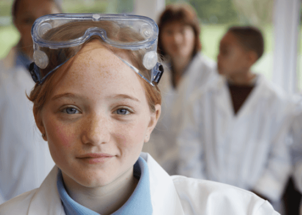 A young scientist, standing in front of three other scientists, wearing a lab coat and goggles on her head.