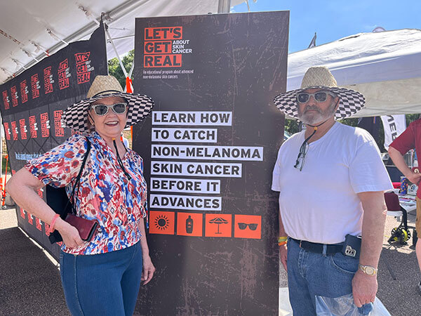  Andre and another man posing next to a Let's Get Real About Skin Cancer branded sign that reads 'Learn how to catch nonmelanoma skin cancer before it advances'.